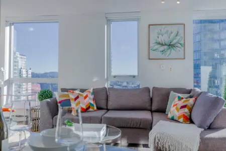 A sunlit living area with floor-to-ceiling windows invites in sweeping city and mountain views, creating a bright, uplifting space to relax. The cozy sectional and vibrant accents make it feel comfortably lived-in, perfect for settling into your Vancouver stay.