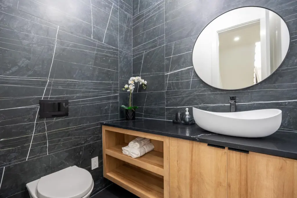This stylish main-floor powder room features striking dark tile walls, a modern vessel sink, and natural wood cabinetry — a sleek and elegant space that adds a touch of sophistication to the home’s design.