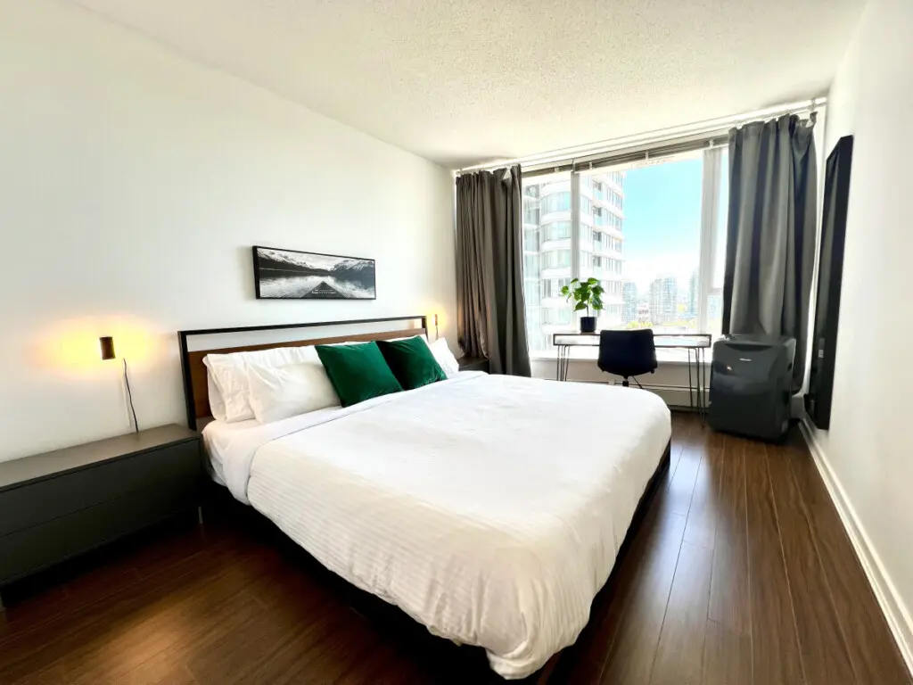 Unwind in the serene comfort of the primary bedroom, featuring a plush king bed, warm lighting, and floor-to-ceiling windows with city views. A peaceful space designed for rest, relaxation, and quiet moments after a day exploring Vancouver.