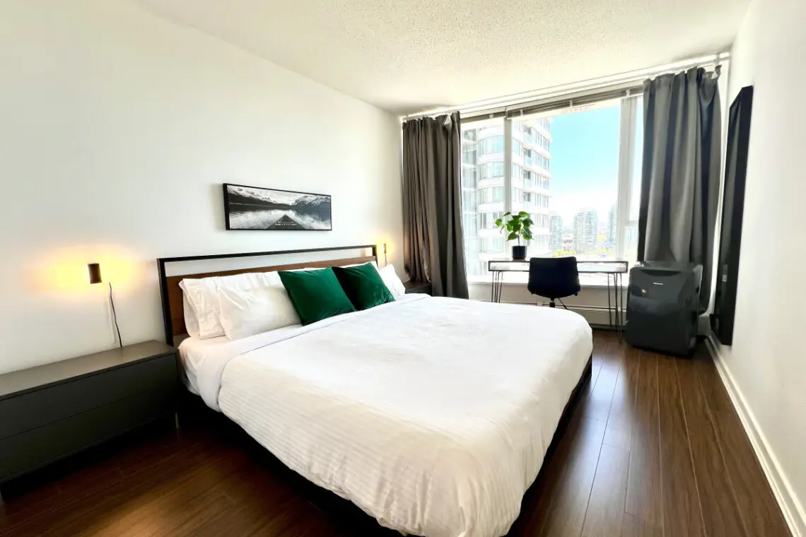 Unwind in the serene comfort of the primary bedroom, featuring a plush king bed, warm lighting, and floor-to-ceiling windows with city views. A peaceful space designed for rest, relaxation, and quiet moments after a day exploring Vancouver.