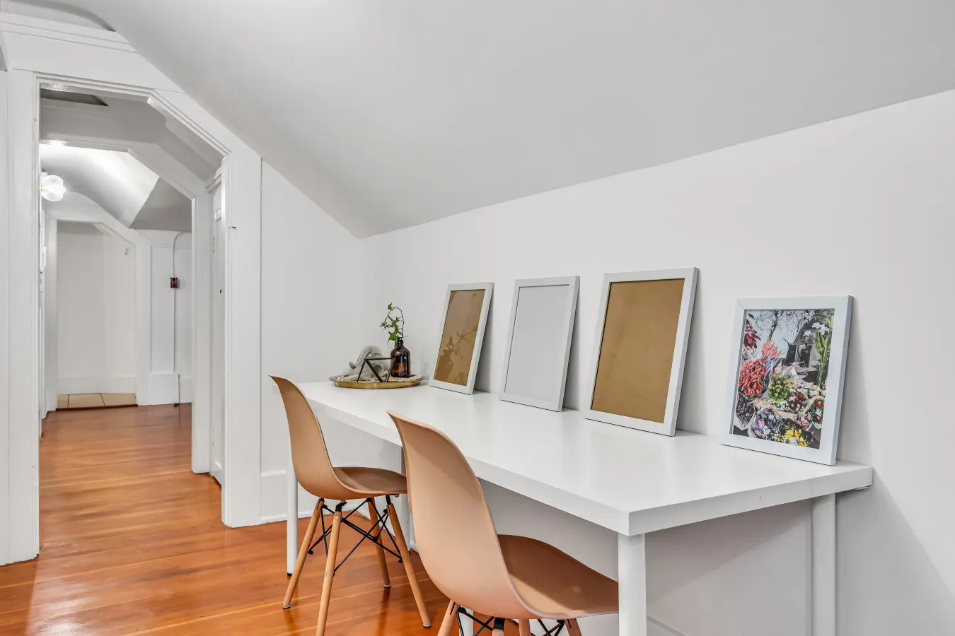 A clean, minimalist workspace featuring a white desk, two chairs, and framed artwork, offering a perfect spot for productivity or creative inspiration.