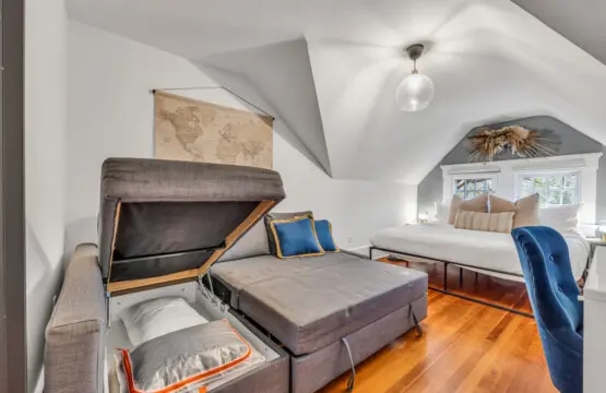 Cozy loft-style bedroom with a plush bed, convertible sofa with hidden storage, and warm natural light streaming through charming window details.