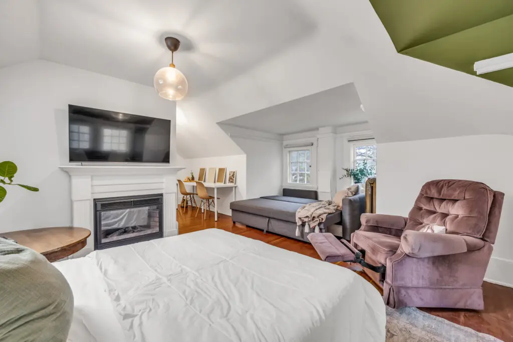 A cozy, well-lit studio featuring a fireplace, a spacious bed, a comfortable recliner, and a dedicated workspace, perfect for both relaxation and productivity.