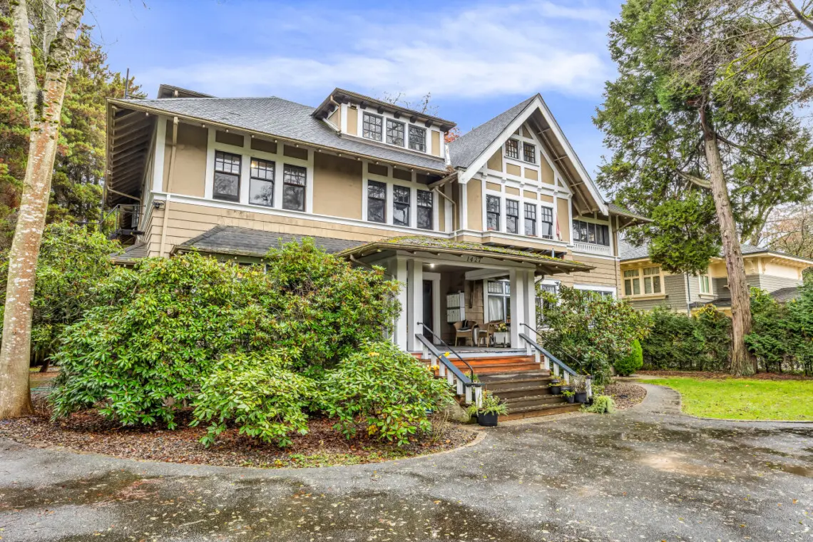 A charming, well-maintained exterior of a large, historic house surrounded by lush greenery, offering a welcoming atmosphere with a front porch and beautifully landscaped yard.
