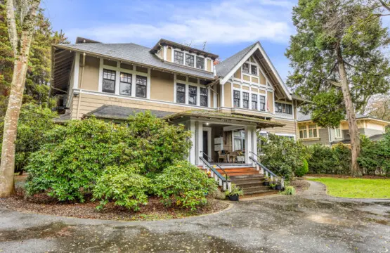 A charming, well-maintained exterior of a large, historic house surrounded by lush greenery, offering a welcoming atmosphere with a front porch and beautifully landscaped yard.
