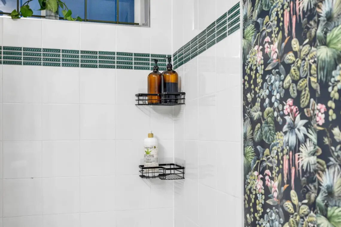 The shower area features green accent tiles and black wire shelves holding bath products, while the backdrop showcases vibrant, nature-inspired wallpaper.