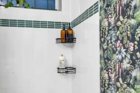 The shower area features green accent tiles and black wire shelves holding bath products, while the backdrop showcases vibrant, nature-inspired wallpaper.