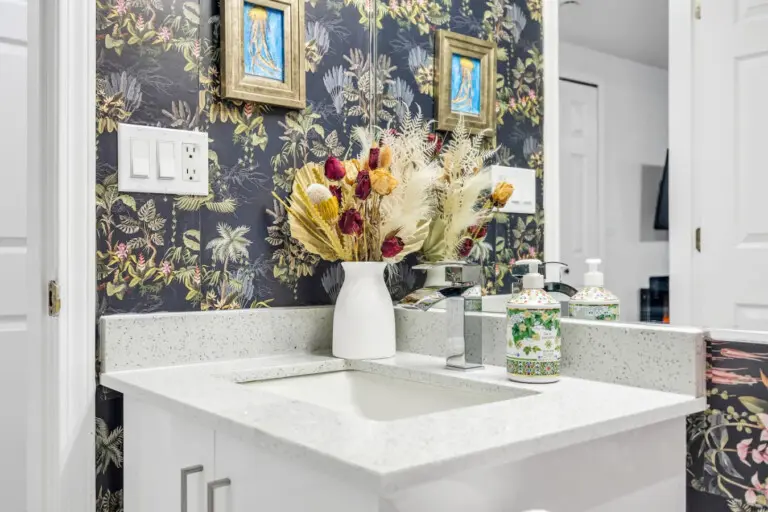 A chic bathroom with a modern sink, floral wallpaper, and decorative accents. The space features a stylish mirror, elegant soap dispenser, and a vase with dried flowers.