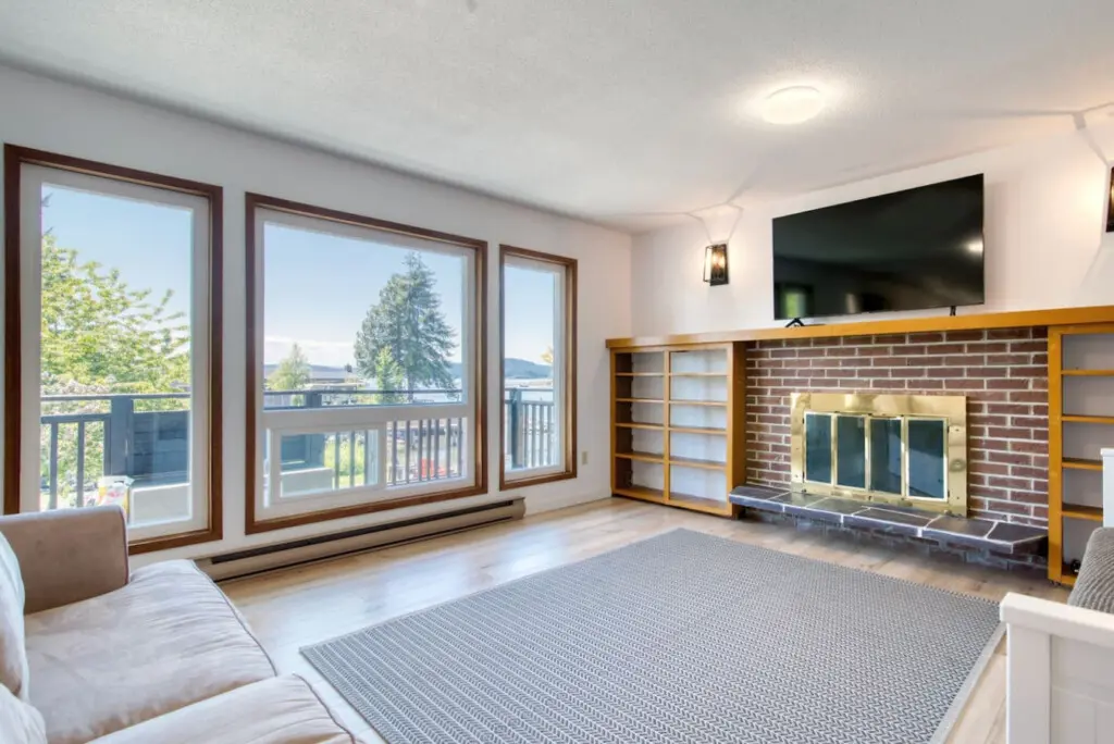 Bright living room with a comfortable sofa, large windows showcasing waterfront views, and a brick fireplace with built-in shelving for cozy gatherings.