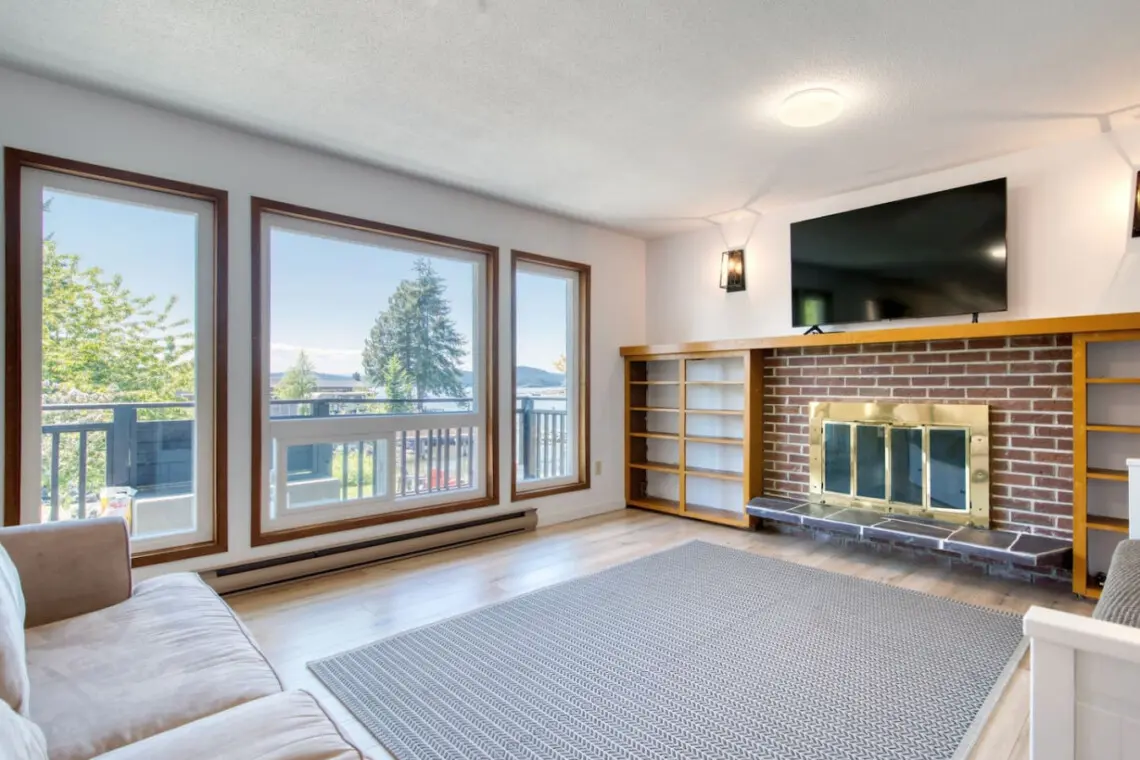 Bright living room with a comfortable sofa, large windows showcasing waterfront views, and a brick fireplace with built-in shelving for cozy gatherings.