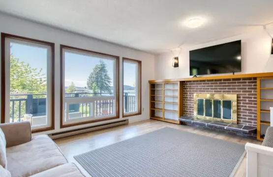 Bright living room with a comfortable sofa, large windows showcasing waterfront views, and a brick fireplace with built-in shelving for cozy gatherings.