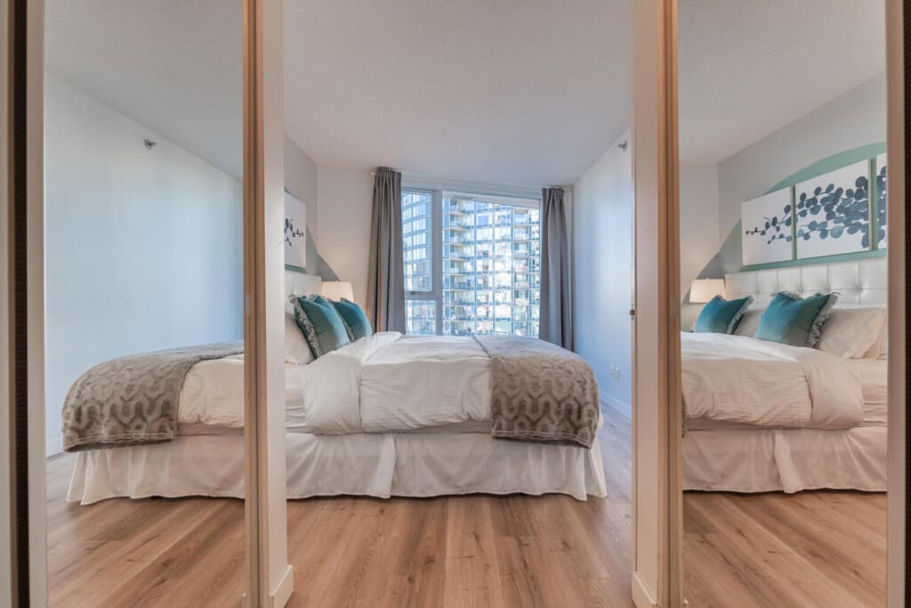 A serene bedroom with mirrored closets enhances both light and space, while soft textures and calming tones create a restful, lived-in feel. Large windows frame city views, offering a bright and comfortable retreat for longer stays.